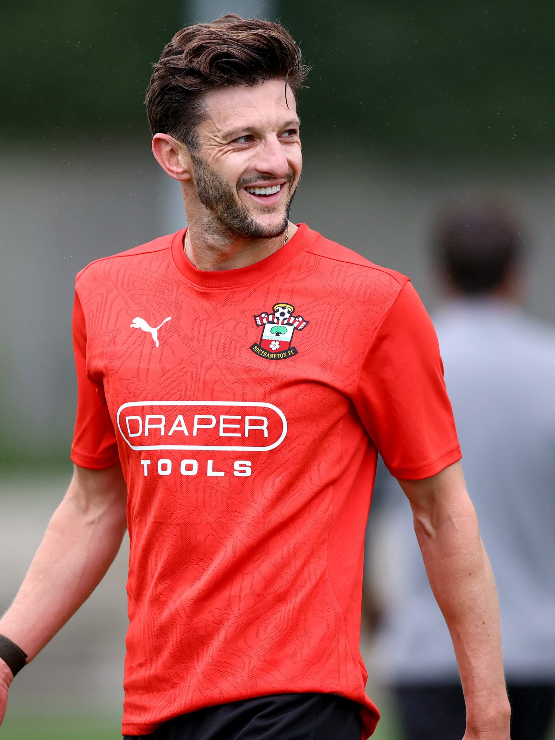 Adam Lallana smiles during training, wearing a red Puma training top.